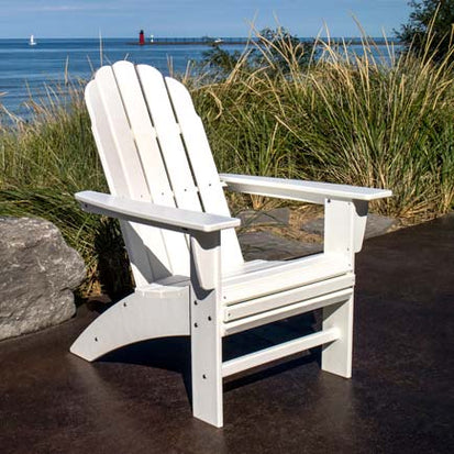 A white Adirondack chair is placed on a patio overlooking a sandy beach and blue sea. Tall grasses and large rocks are nearby, and a red lighthouse is visible in the distance. The sky is clear and sunny, creating a serene coastal scene.