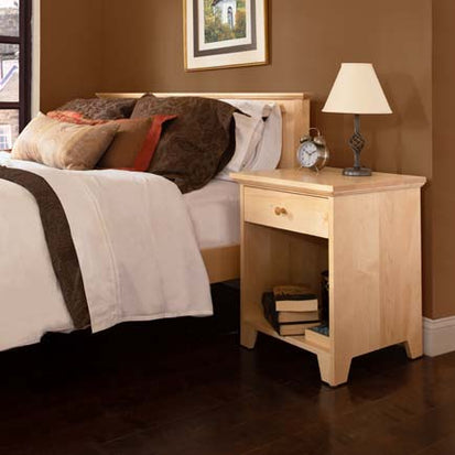 A bedroom featuring a wooden bed with white and brown bedding, a wooden nightstand with an alarm clock, a stack of books, and a lamp. The walls are painted brown, and there is a framed picture above the bed. Dark wooden flooring is visible.