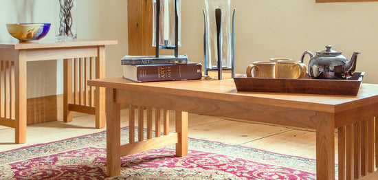 A cozy living room features two wooden tables with slatted sides. The foreground table holds books, a tea set, and a decorative tray, while the background table displays a bowl. Both tables rest atop a red patterned rug on a wooden floor.