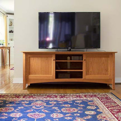 A flat-screen TV sits on a wooden entertainment center with two closed cabinets and central open shelving. The unit is placed against a beige wall, and a blue area rug with a red and yellow pattern covers the wooden floor in front of it.