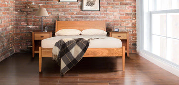 This image shows a bedroom with a wooden bed against a brick wall. The bed has beige bedding and a plaid blanket. Two wooden nightstands with lamps are on either side of the bed, and a window is on the right. Framed pictures are hung above the bed.