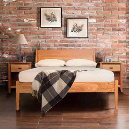 A bedroom features a wooden bed with a white mattress and two pillows. A black and white checkered blanket is draped across the foot of the bed. A bedside table with a lamp is on each side. A brick wall in the background has two framed artworks hanging above the bed.