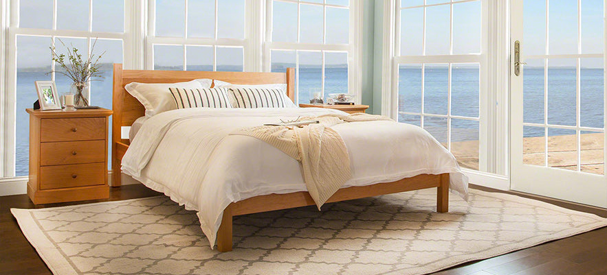 A bedroom with a wooden bed frame and white bedding set on a patterned rug. Two wooden nightstands flank the bed, each with a plant and framed photo. Large windows offer an ocean view, and a wooden door provides access to the outside.