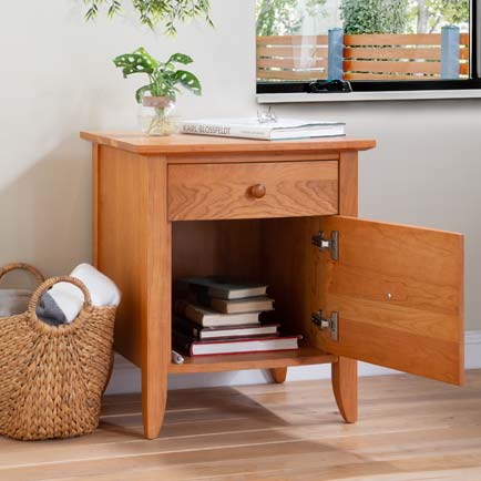 A wooden nightstand with a drawer and an open lower cabinet containing books. A small plant in a vase sits on top of the nightstand. There is a wicker basket with blankets next to the nightstand, and a window in the background shows an outdoor space.
