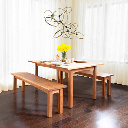 A dining area with a wooden table set for two, featuring plates, cups, and breakfast items. Two wooden benches are on either side of the table. A bunch of yellow daffodils in a vase is centered on the table, and an abstract chandelier hangs above.