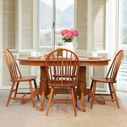 A wooden dining table with four matching chairs is set with plates, cups, and a white pitcher containing pink flowers. The table is situated in a sunlit room with large windows and white shutters, offering a view of the outdoors.