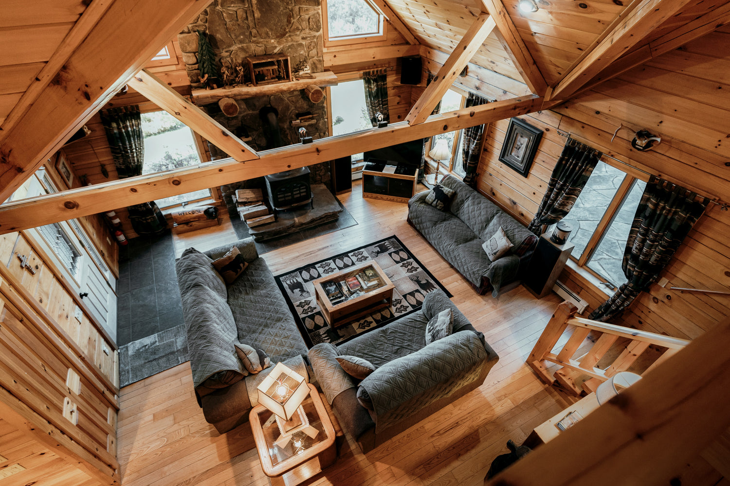 A cozy living room in a wooden cabin viewed from an elevated perspective. The room features a stone fireplace, two gray sofas, a patterned rug, a wooden coffee table, and various decorative items. Large windows and exposed beams contribute to the rustic ambiance.