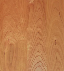 A close-up view of a wooden surface displaying a natural grain pattern with wavy lines and occasional knots. The wood has a light to medium brown hue, giving it a warm and rustic appearance.