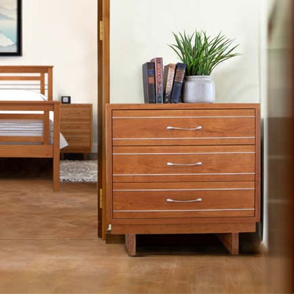 A wooden dresser with three drawers, a potted plant, and several books on top is set in a bedroom. The room also features a wooden bed frame and a nightstand in the background, with light-colored walls and wooden flooring.