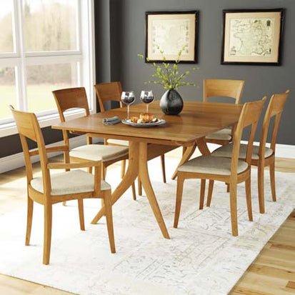 A wooden dining table is set with two glasses of wine, a plate, and utensils. Six wooden chairs with cushioned seats surround the table. The room has dark gray walls with two framed maps and large windows. A rug is under the table and chairs.