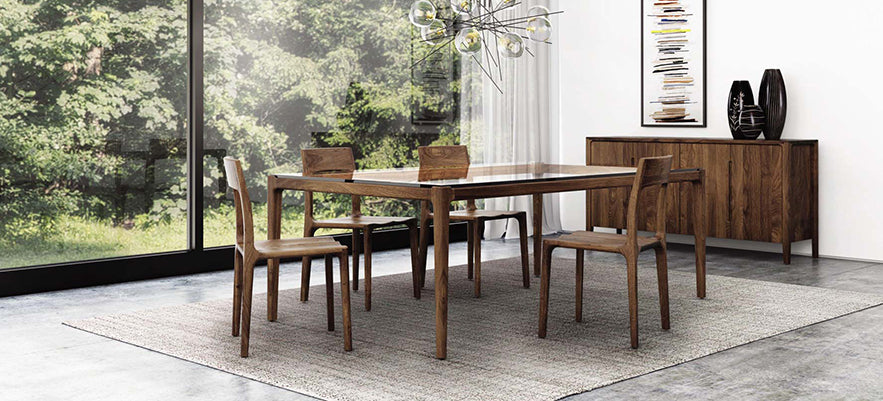 A dining room features a wooden table with four matching chairs on a grey rug. Behind the table is a wooden sideboard with vases and a framed artwork. Large windows provide a view of lush green trees, and a modern chandelier hangs above the table.