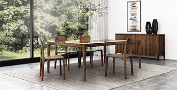 A dining room features a wooden table with four matching chairs on a grey rug. Behind the table is a wooden sideboard with vases and a framed artwork. Large windows provide a view of lush green trees, and a modern chandelier hangs above the table.