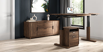 A modern office space features a wooden sit-stand desk with an open laptop, a two-drawer pedestal, a wooden sideboard, and a decorative wall piece. The room has large windows, light wood flooring, and black vertical accent paneling on one wall.