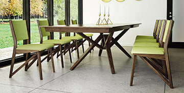 A modern dining room features a wooden table with cross-leg design and six green upholstered chairs. Three gold candlesticks with white candles are placed in the center of the table. Large windows on the left side provide a view of an outdoor area with trees.