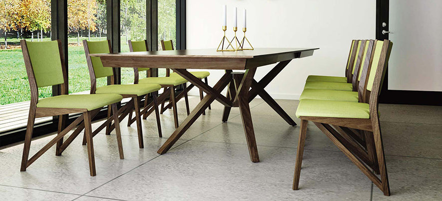 A modern dining room features a wooden table with cross-leg design and six green upholstered chairs. Three gold candlesticks with white candles are placed in the center of the table. Large windows on the left side provide a view of an outdoor area with trees.