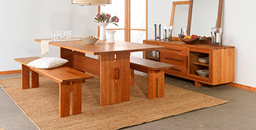 A dining room with a wooden table, two benches, and a chair. The table has a white bowl and a vase with flowers. A matching wooden sideboard with decor items, including a mirror, is against the wall. The room features a natural fiber rug and neutral decor.