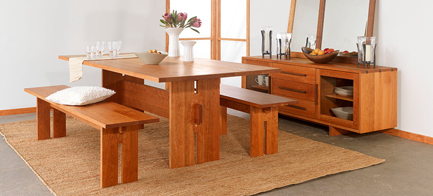 A wooden dining table set with two benches is placed on a light brown rug. The table has a white vase with flowers and bowls. Behind it is a matching wooden sideboard with decor items, including a mirror, candlesticks, and bowls of fruit.