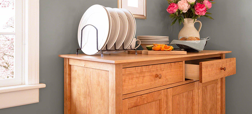 Wooden sideboard with an open drawer, featuring a rack holding plates and topped with a vase of flowers and bowl of oranges.