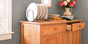 Wooden sideboard with an open drawer, featuring a rack holding plates and topped with a vase of flowers and bowl of oranges.