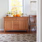 A Vermont Furniture Designs Heartwood Shaker Sideboard made of solid hardwoods is situated near a window, adorned with pumpkins and a vase of flowers, accompanied by a wooden chair; a patterned rug lies beneath, and a