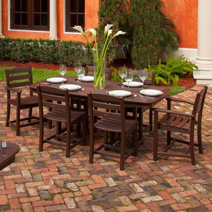 A rectangular wooden outdoor dining table is set with plates, wine glasses, and a vase of white flowers. The table is surrounded by six matching wooden chairs. The setup is on a brick patio with greenery and an orange building in the background.