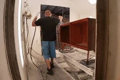 A man in a black shirt and blue shorts is spray painting a large piece of furniture in a spray booth. The furniture appears to be a cabinet, and it is being coated with a reddish hue. The setting looks industrial, with metal grates on the floor.