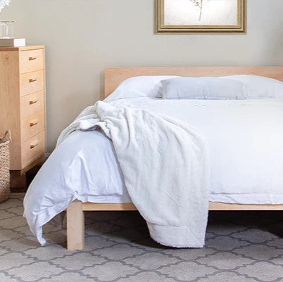 A neatly made bed with a light wooden frame is positioned against a wall in a bedroom. The bed is covered with a white comforter and has two pillows. A white blanket is casually draped over the side. There is a wooden dresser with drawers to the left.