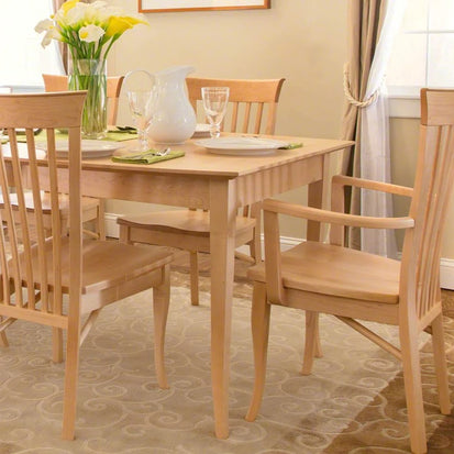 A dining area with a light wooden table and matching wooden chairs. The table is set with plates, glasses, green napkins, and a white pitcher. A vase with white and yellow flowers sits at the back. The room has beige walls, a window with curtains, and a patterned rug.