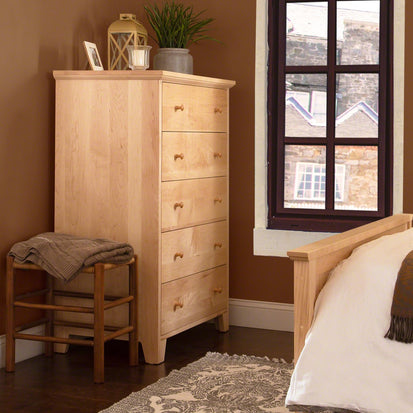 A wooden dresser with six drawers stands beside a window in a bedroom with brown walls. A plant, lantern, and candle sit on top of the dresser. A wooden chair with a folded blanket is next to the dresser, and a bed with white linens is partially visible.