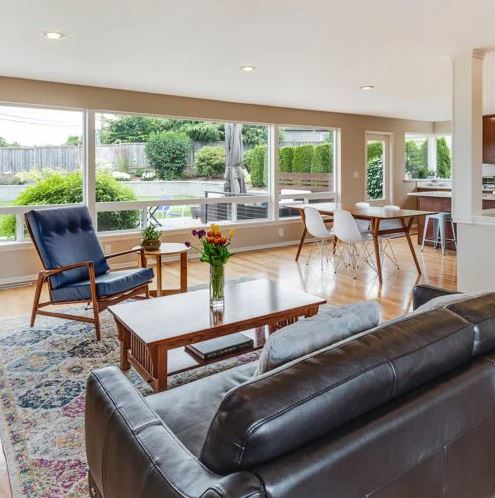 A modern living room with large windows and wooden flooring. It features a black leather sofa, a wooden coffee table with a vase of flowers, a blue armchair, and a patterned rug. In the background, there is a dining area with a table and white chairs.
