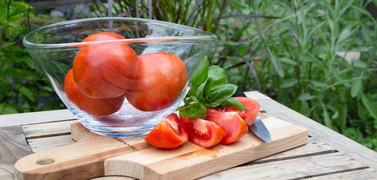 A clear glass bowl with whole tomatoes sits on a wooden surface next to a cutting board. On the cutting board are sliced tomatoes, a few basil leaves, and a small knife. The background shows green foliage.