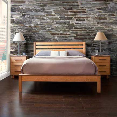 A bedroom with a stone accent wall, a wooden bed frame, and a gray comforter. Two wooden nightstands with drawers are on each side of the bed, each with a lamp. The room has dark wood flooring and a large window letting in natural light.