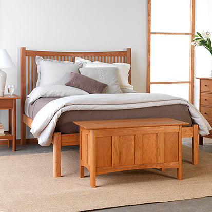 A bedroom featuring a wooden bed frame with white and grey bedding, a wooden blanket chest at the foot of the bed, a wooden nightstand with a white lampshade, and a flower vase on a wooden drawer chest against a white wall. A beige rug covers the floor.