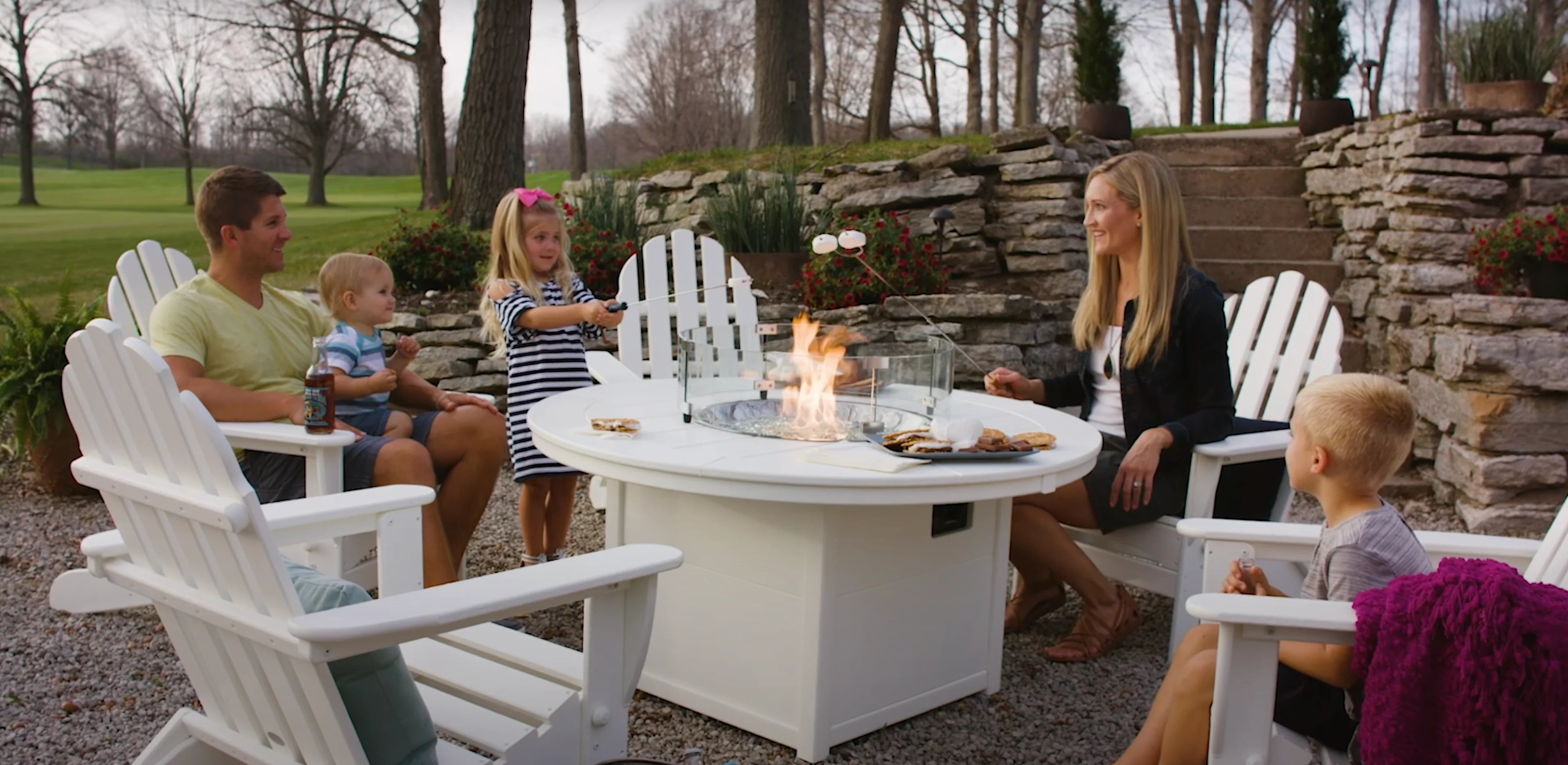 A group of six people, including three adults and three children, sit around a white outdoor fire pit table, surrounded by white Adirondack chairs. The setting is a well-manicured backyard with a stone retaining wall and trees in the background.