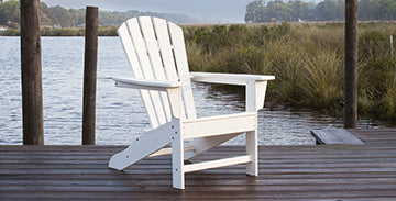 A white Adirondack chair is placed on a wooden dock by a body of water. The background includes grassy marshland and several wooden posts. The scene appears calm and serene.