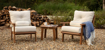 Two cushioned outdoor chairs with wooden frames face each other with a small wooden table in between. The table holds a tray with two cups, a teapot, and a glass. A stack of firewood is in the background, and a blue throw is draped over one chair.
