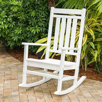 A white wooden rocking chair sits on a paved patio area. It is positioned near green shrubs and palm plants, creating a serene outdoor setting. The chair has a classic slatted backrest and armrests, designed for comfort and relaxation.