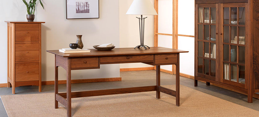 A wooden desk with three drawers is placed on a beige rug. On the desk are a lamp, books, and a ceramic vase. Behind the desk, there is a tall wooden dresser and a wooden cabinet with glass doors. A framed picture hangs on the wall.