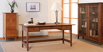 A wooden study room features a desk with drawers, a table lamp, and decorative items. The room includes a tall chest of drawers on the left, a glass-fronted bookcase on the right, and a framed photo on the wall. A beige rug covers the floor.