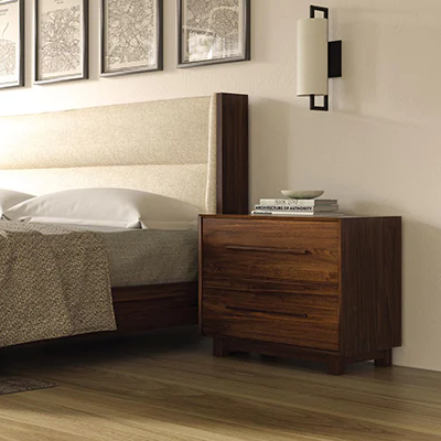 A modern bedroom features a wooden bed with a cushioned headboard and a matching wooden nightstand with three drawers. On the nightstand are a few books and a small plate. Above the nightstand, a black and white wall-mounted light fixture is visible.