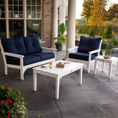 Outdoor patio set with a white frame and navy-blue cushions, consisting of a loveseat, chair, coffee table, and side table. The table holds drinks and small plates. The patio is furnished with potted plants and overlooks a garden and trees in autumn colors.