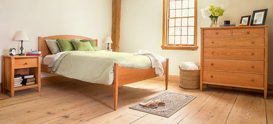 A roomy bedroom with a hardwood floor, a wooden bed frame with a green and white bedspread, a wooden nightstand with a lamp and books, a wooden dresser with framed photos and a vase of flowers on top, a wicker basket, and a window with natural light.
