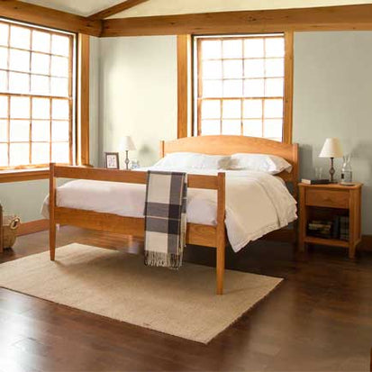 A bedroom with wooden furniture including a bed, two nightstands, and two lamps. The bed is made with white sheets and has a plaid blanket draped over the footboard. Two large windows with wooden frames allow natural light to fill the room. The floor is hardwood.