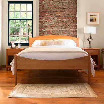 A bedroom features a wooden bed with white bedding, flanked by two matching wooden nightstands. A window with black trim is on the left, and a brick accent wall is behind the bed. A framed picture and a table lamp are on the right nightstand. A rug is on the wooden floor.